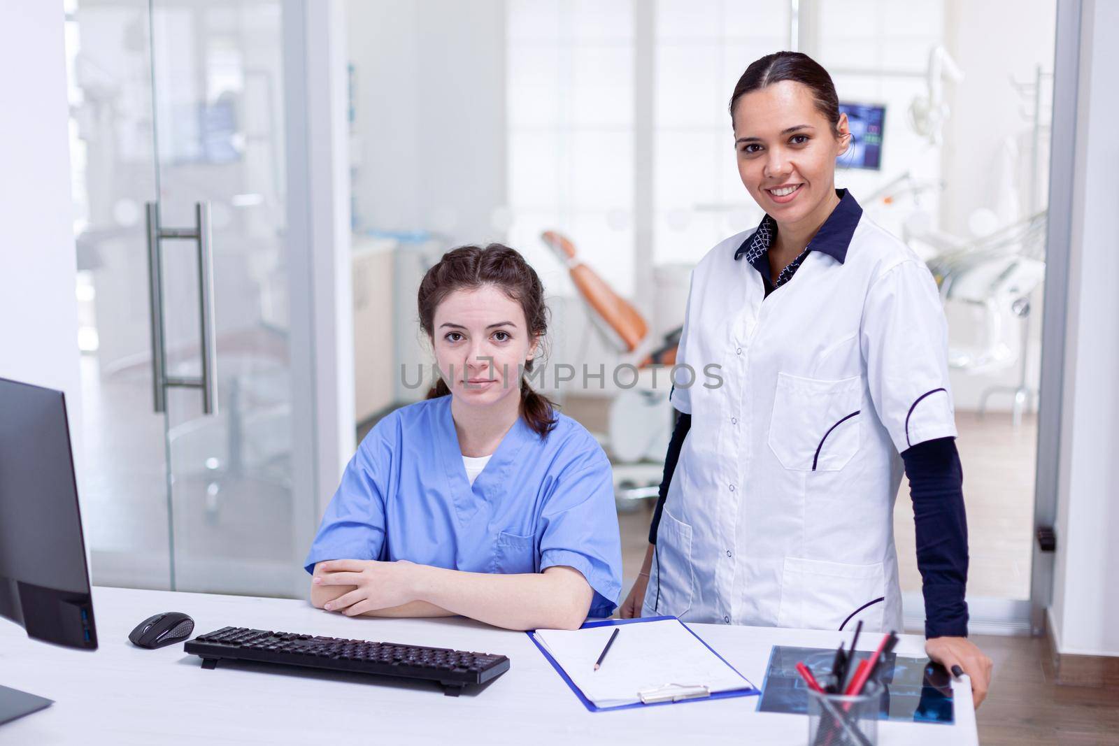 Medical team in dental office smiling at camera wearing uniform. Stomatology assistant and teeth doctor discussing in reception of dental office