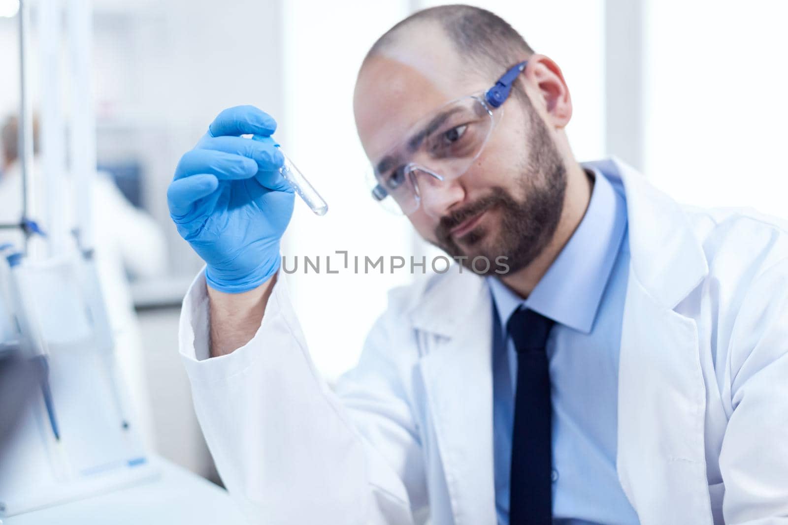 Doctor scientist in professional lab looking pensive at test tube with chemical substance. Researcher in biotechnology sterile lab holding analysis in tube wearing gloves and protection glasses.