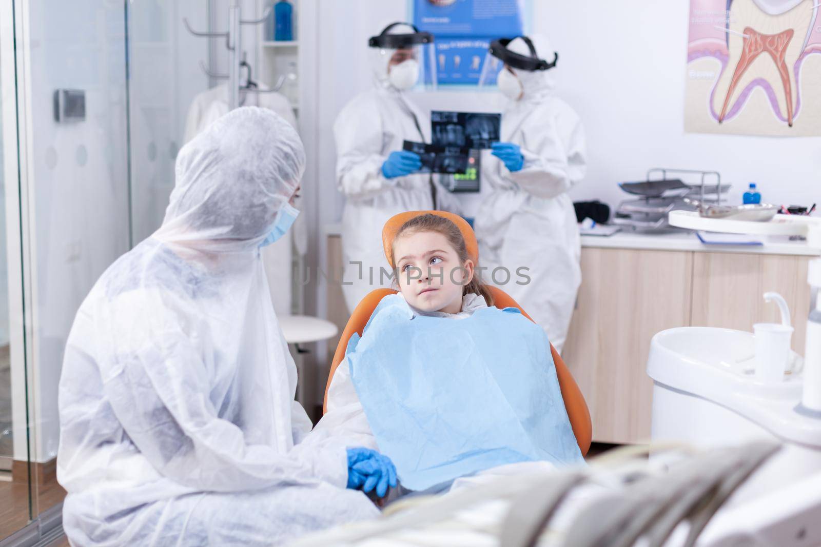 Girl looking pensive mother sitting on dental chair wearing coverall because of coronavirus outbreak. Stomatologist during covid19 wearing ppe suit doing teeth procedure of child sitting on chair.
