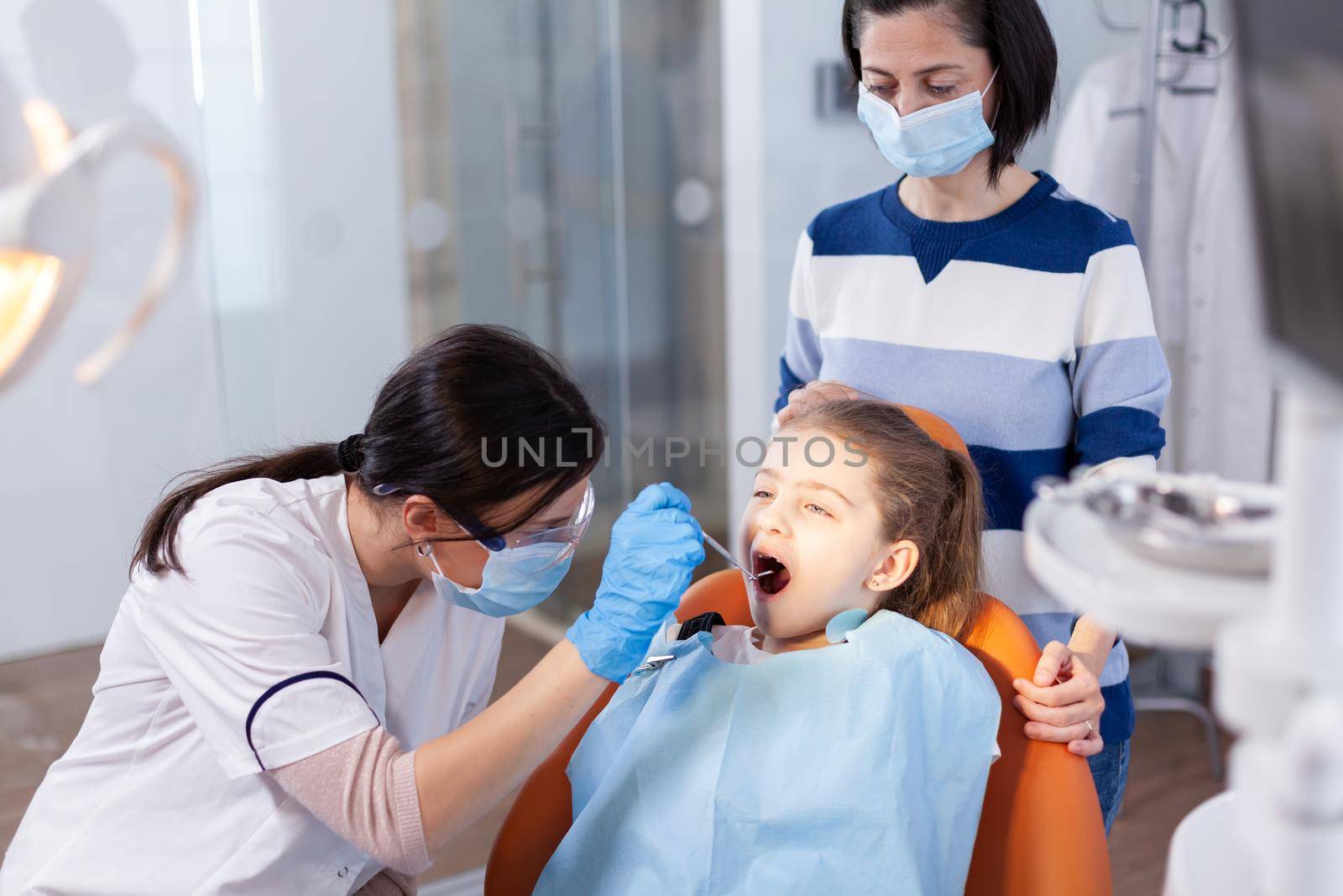 Stomatolog using angled mirror during oral examining of girl sitting on dental chair. Dentistry specialist during child cavity consultation in stomatology office using modern technology.