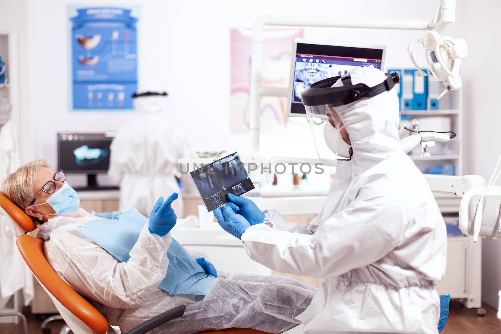 Stomatolog in safety gear agasint coronavirus holding x-ray pf senior patient. Elderly woman in protective uniform during medical examination in dental clinic.