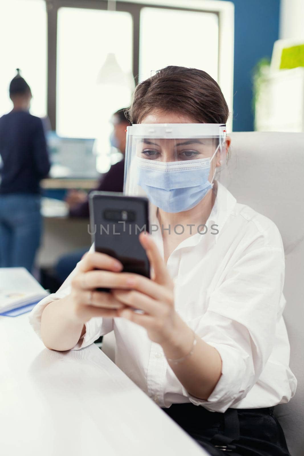 Entrepreneur wearing face mask as safety precaution against covid 19 chatting on smartphone at workplace.Multiethnic coworkers working respecting social distance in financial company. New normal.