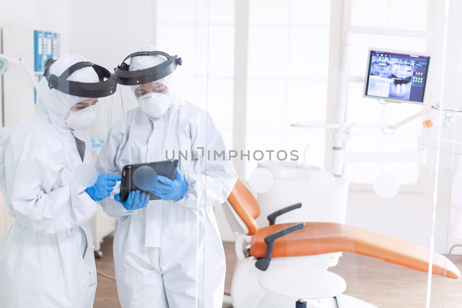 Dentist and nurse during covid-19 outbreak in ppe suit usint tablet pc. Stomatology team in dental office wearing protective suit agasint contagious coronavirus during global pandemic.