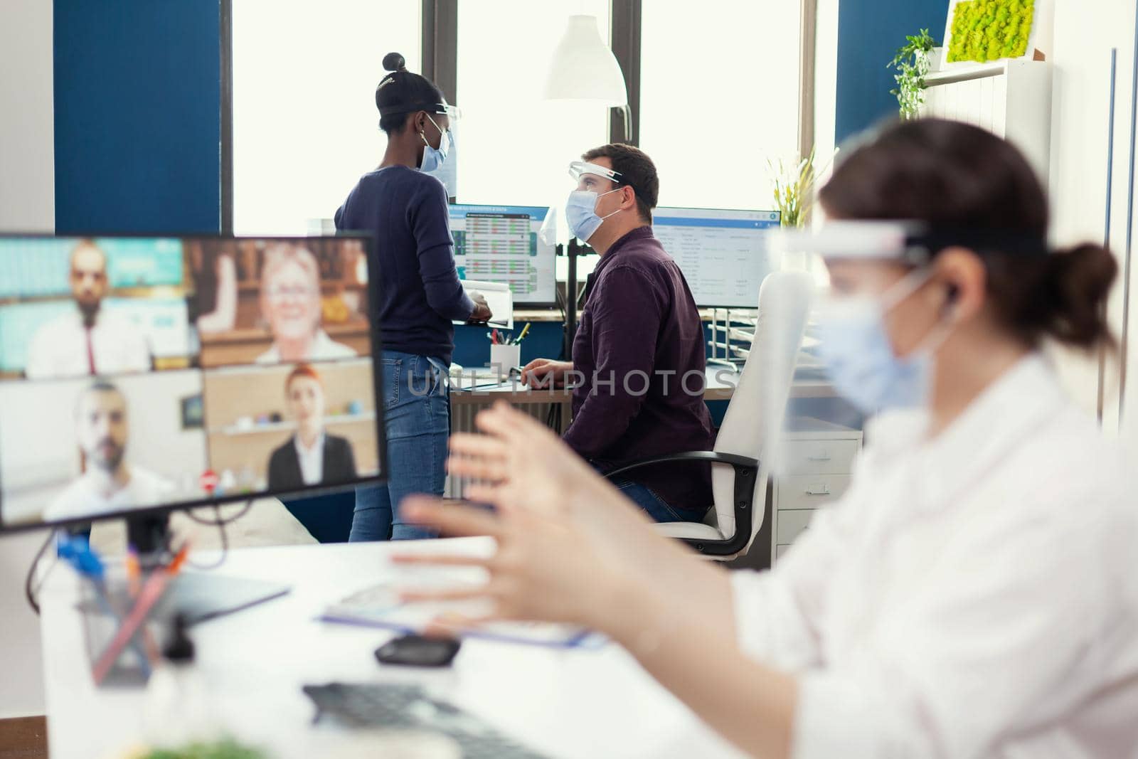 African woman having a conversation with her coworker waring face mask at workplace wearing face mask. Entrepreneur having videocall while colleagues working respecting social distance during global pandemic.