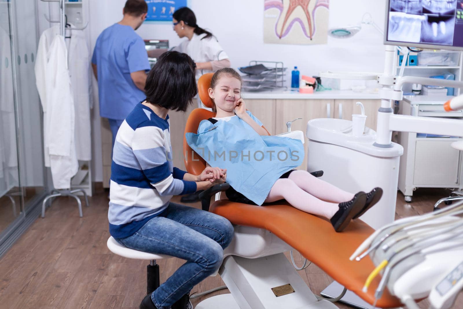 Little girl with painful expression because of thoot cavity waiting for treatment from professiona dentist. Child with her mother during teeth check up with stomatolog sitting on chair.