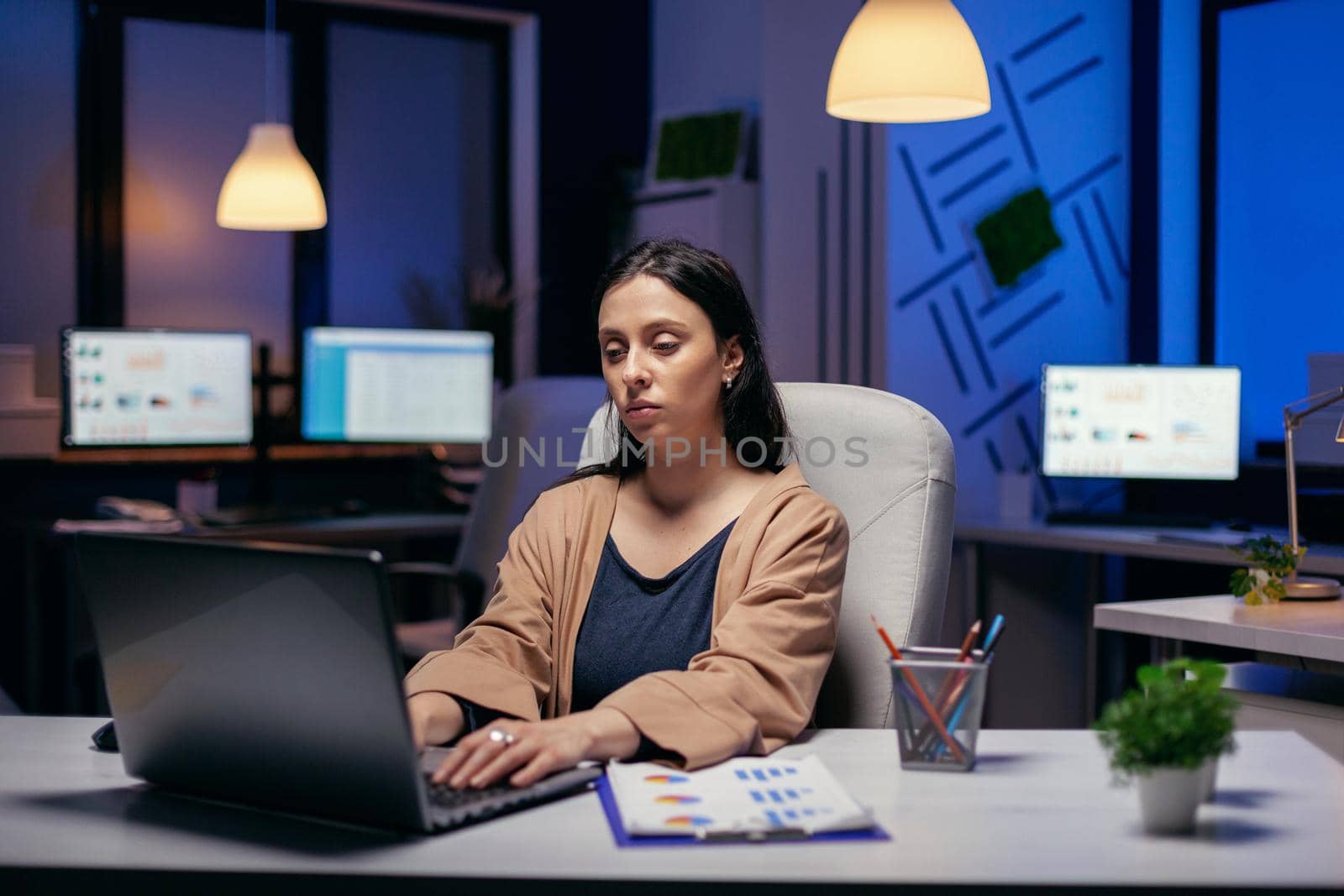 Young woman working on the laptop doing overtime to finish a deadline sitting at her workplace. Smart woman sitting at her workplace in the course of late night hours doing her job.