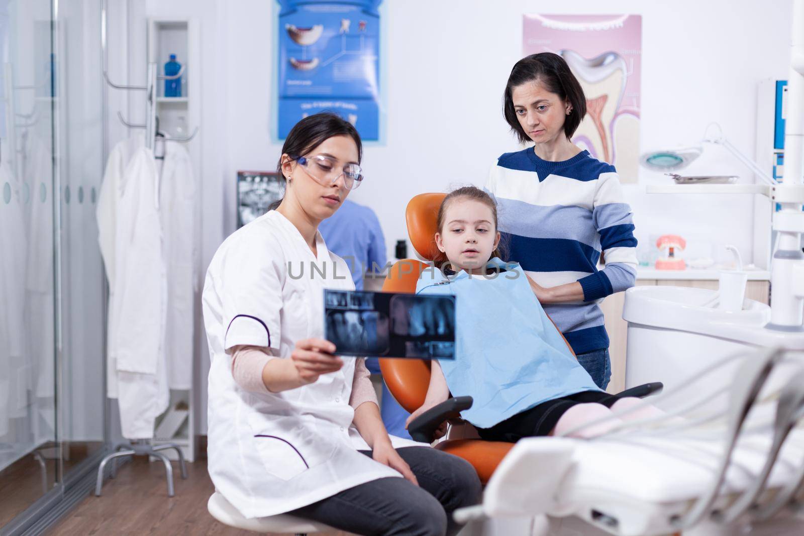 Little girl and parent listening dentist advice pointing at dental radiography. Stomatologist explaining teeth diagnosis to mother of child in health clinic holding x-ray.