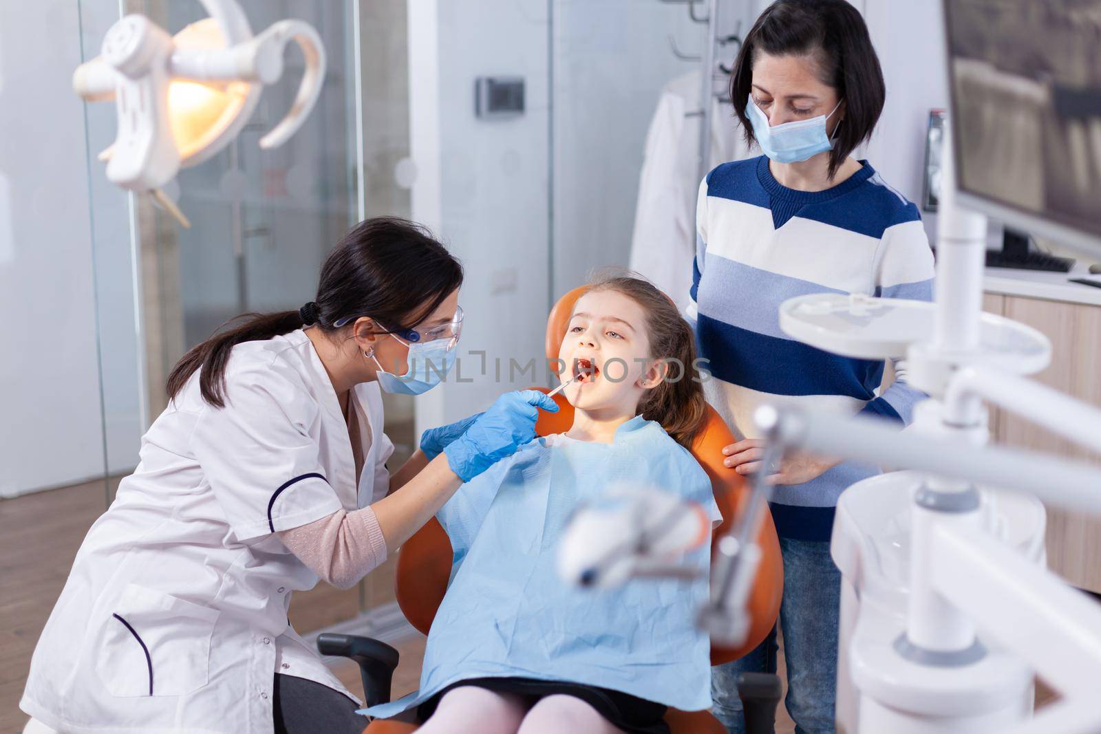 Dentist using mirror in the course of kid mouth examination sitting on dental chair. Dentistry specialist during child cavity consultation in stomatology office using modern technology.