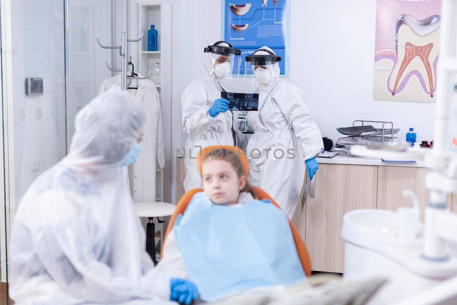 Dentist doctor and assistant holding dental radiography of little kid patient in the course of coronavirus. Stomatologist during covid19 wearing ppe suit doing teeth procedure of child sitting on chair.