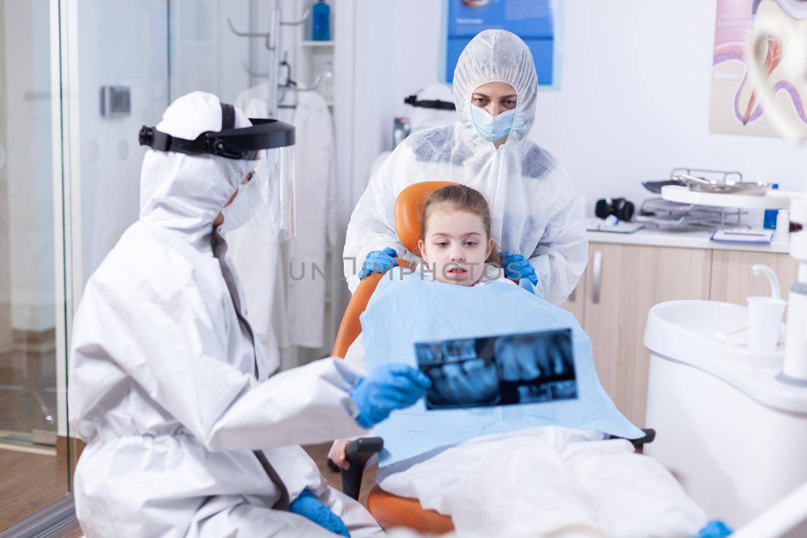 Little girl and mother wearing ppe suit looking at dental radiography in dentist office during covid19 pandemic. Stomatolog in protectie suit for coroanvirus as safety precaution holding child teeth x-ray during consultation.