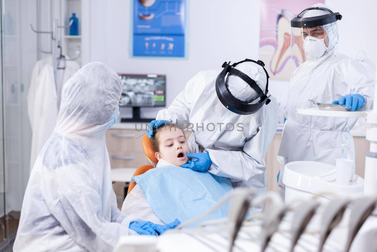 Pediatric stomatolog doing oral hygine procedure on little girl with bib wearing ppe suit. Dentist in coronavirus suit using curved mirror during teeth examination of child.