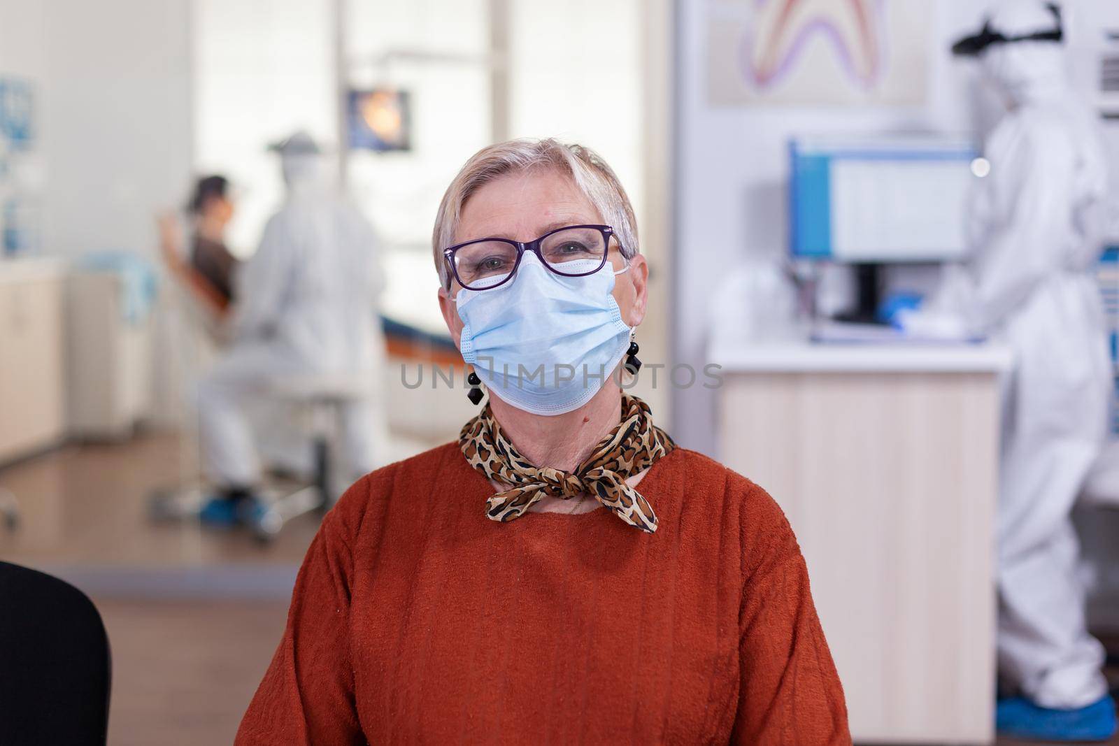 Portrait of retired patient in dental office looking on camera wearing face mask sitting on chair in waiting room clinic while doctor working. Concept of new normal dentist visit in coronavirus outbreak.
