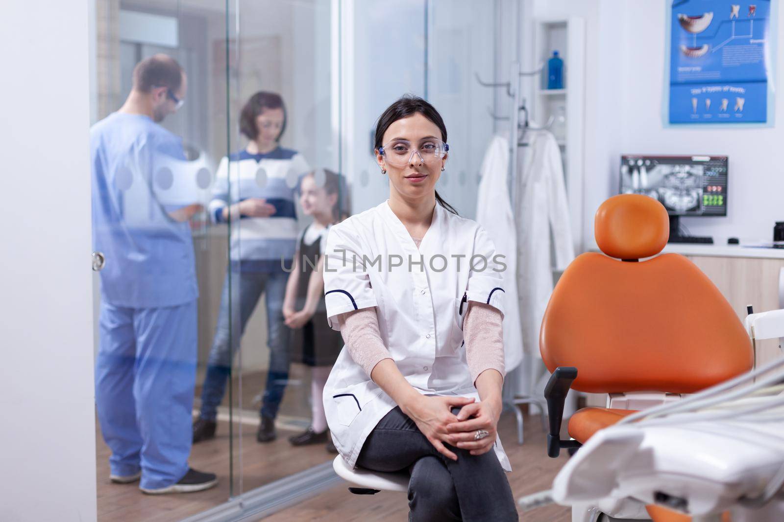 Dentist in dentistiry office sitting on chair while assistant is talking with patients by DCStudio
