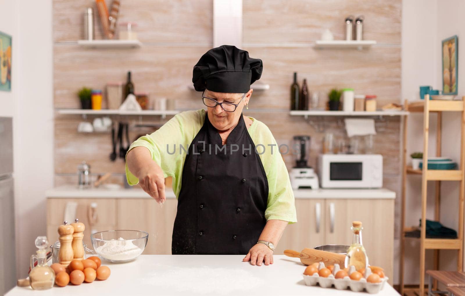 Woman preparing tasty pizza sprinkling baking powder on kitchen table. Happy elderly chef with uniform sprinkling, sieving sifting raw ingredients by hand.