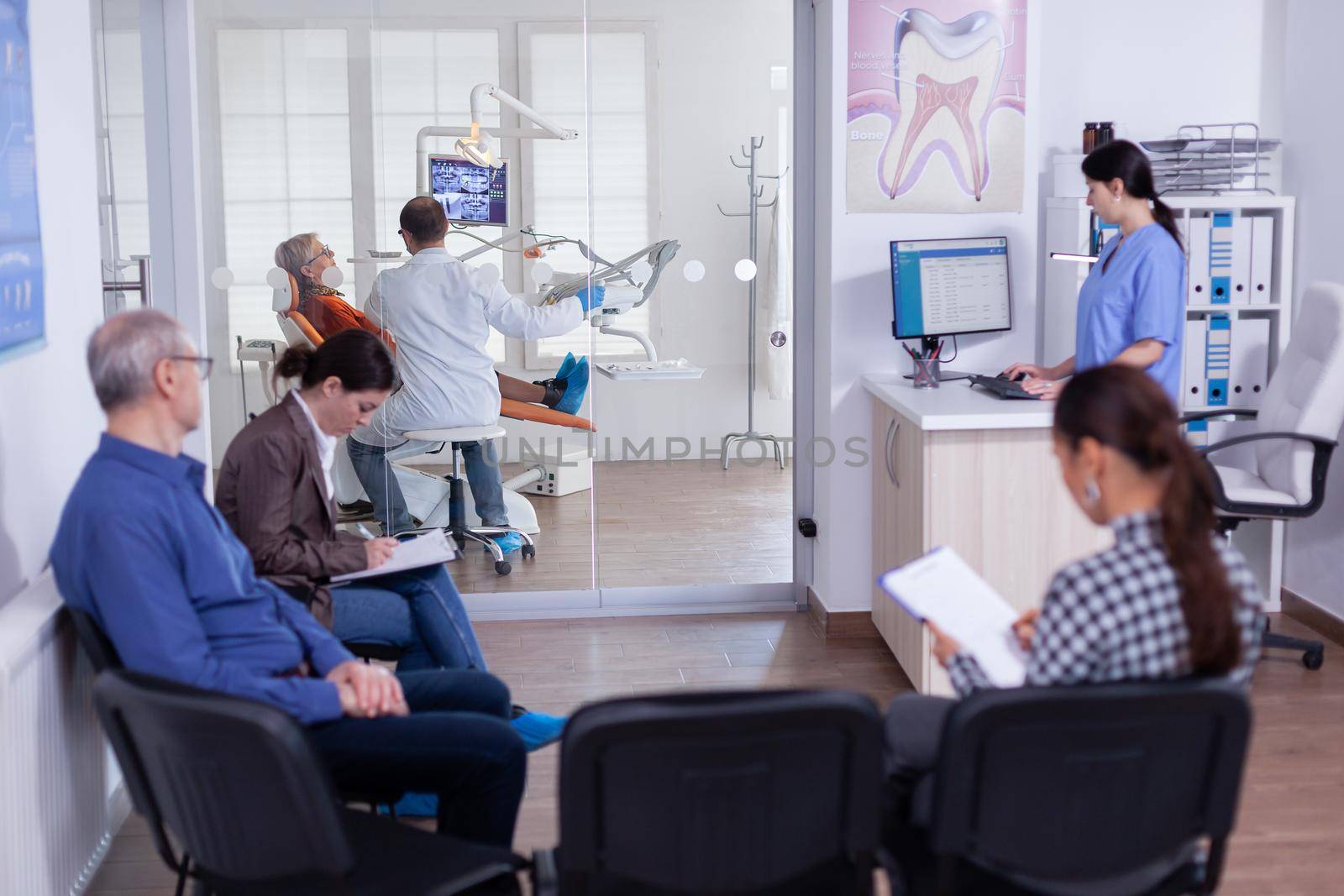 Dentist doctor treating senior woman teeth in stomatology clinic. Patient in waiting area filling dentistiry form for surgery. Assitant working on computer in dental reception.