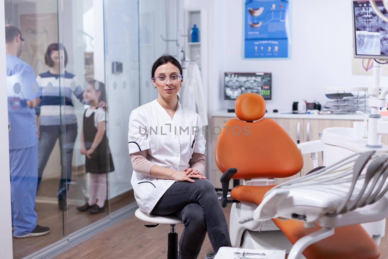 Ortodontist in dentistiry office sitting beside tools tray with assistant discussing with patietns in the background. Stomatolog in professioanl teeth clinic smiling wearing uniform looking at camera.