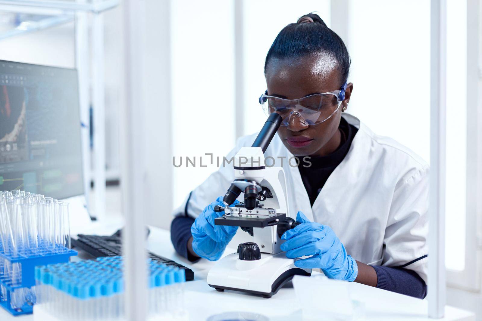 African chemist doing investigation using microscope analysing sample on glass slide. Black healthcare scientist in biochemistry laboratory wearing sterile equipment.