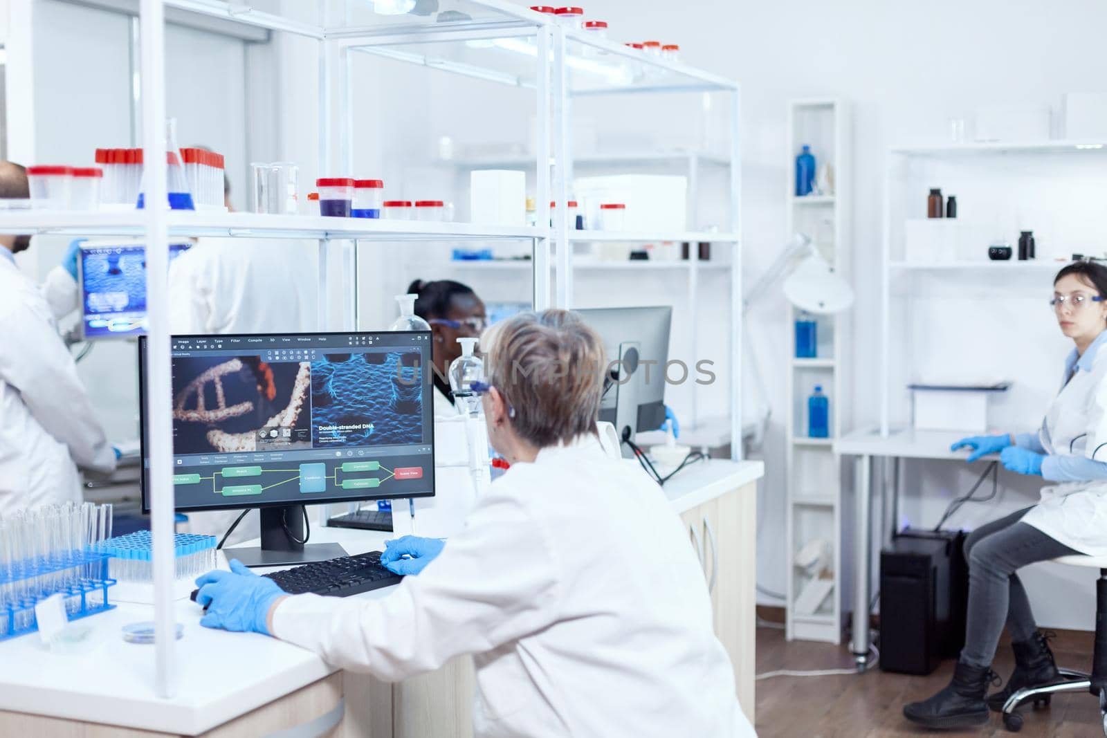Elderly virologist working on computer with virus on display in modern facility. Senior scientist in pharmaceuticals laboratory doing genetic research wearing lab coat with team in the background.