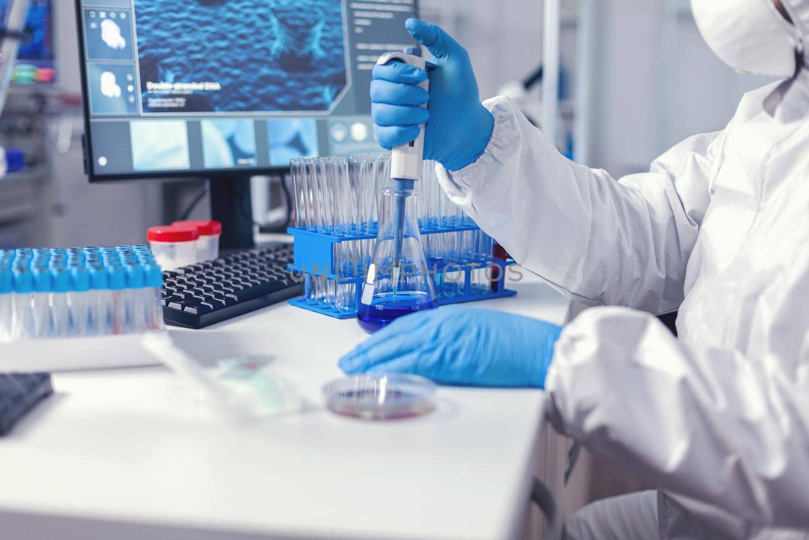 Professional lab scientist dropping sample using automatic pipette dressed in protection suit. Chemist in modern laboratory doing research using dispenser during global epidemic with covid-19.