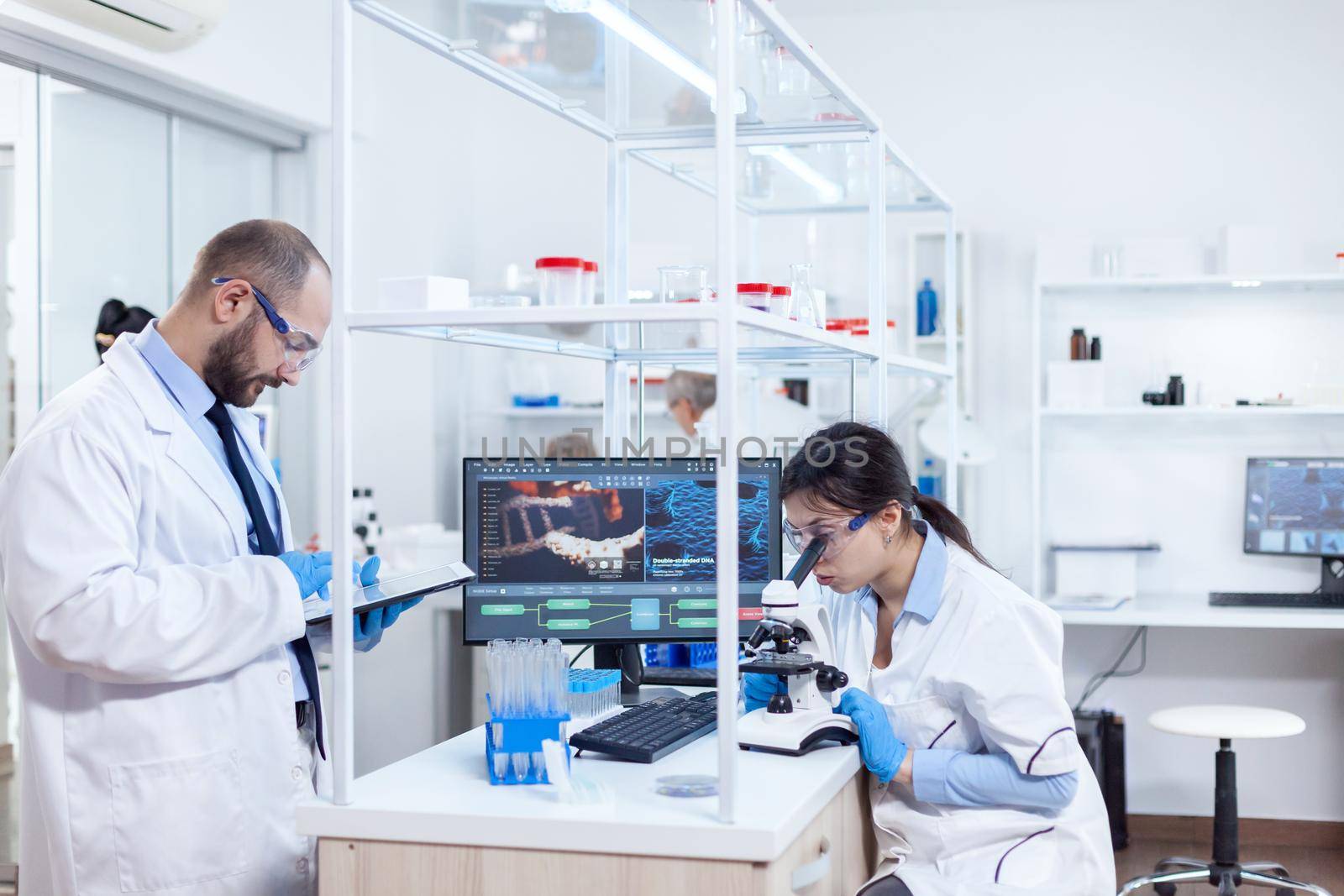 Chemist wearing safety glasses and using tablet pc in lab. Team of researchers doing pharmacology engineering in sterile laboratory for healthcare industry with african assistant in the background.