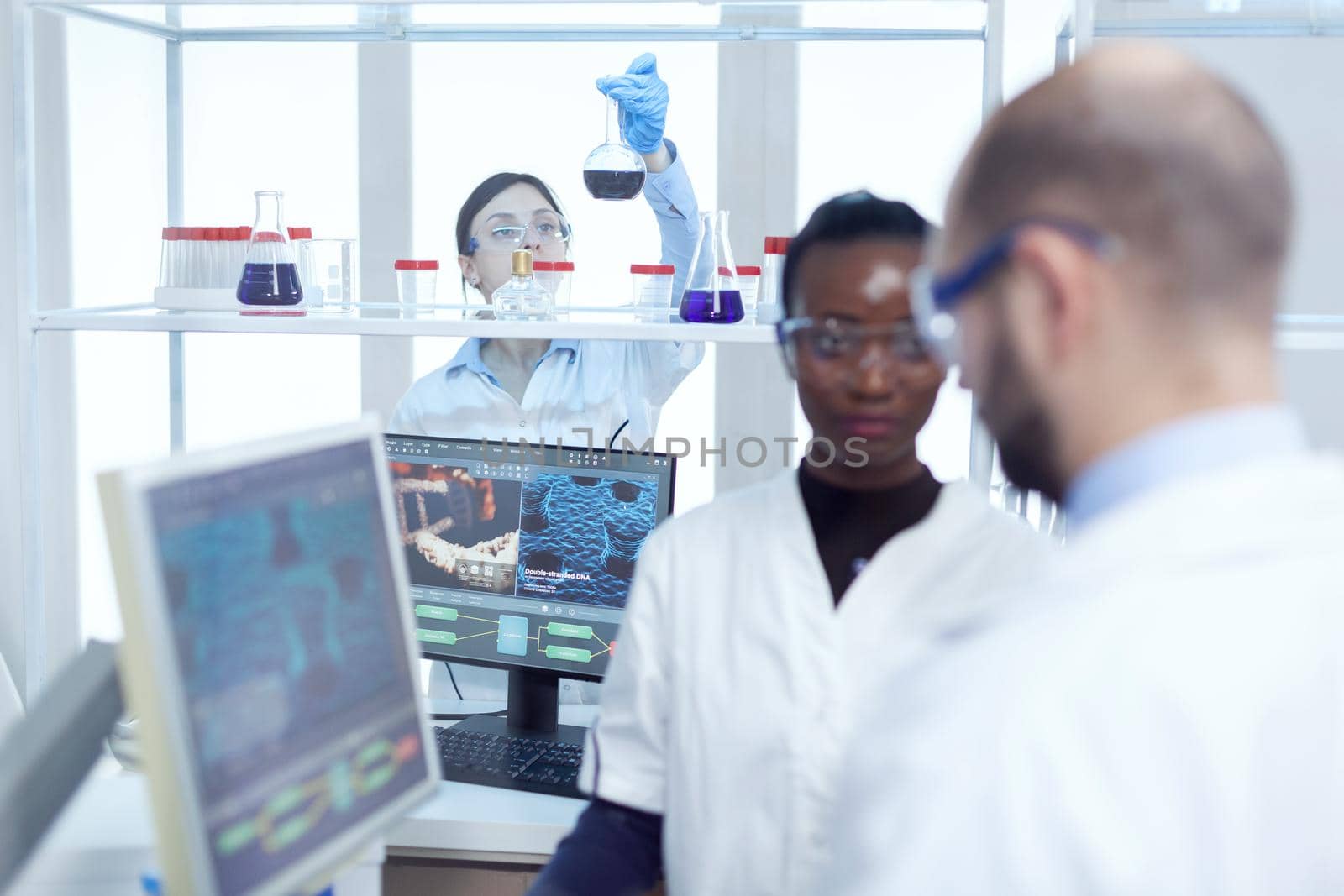 Expert chimist examining liquid in glass recipient. Multiethnic team of medical researchers working together in sterile lab wearing protection glasses and gloves.