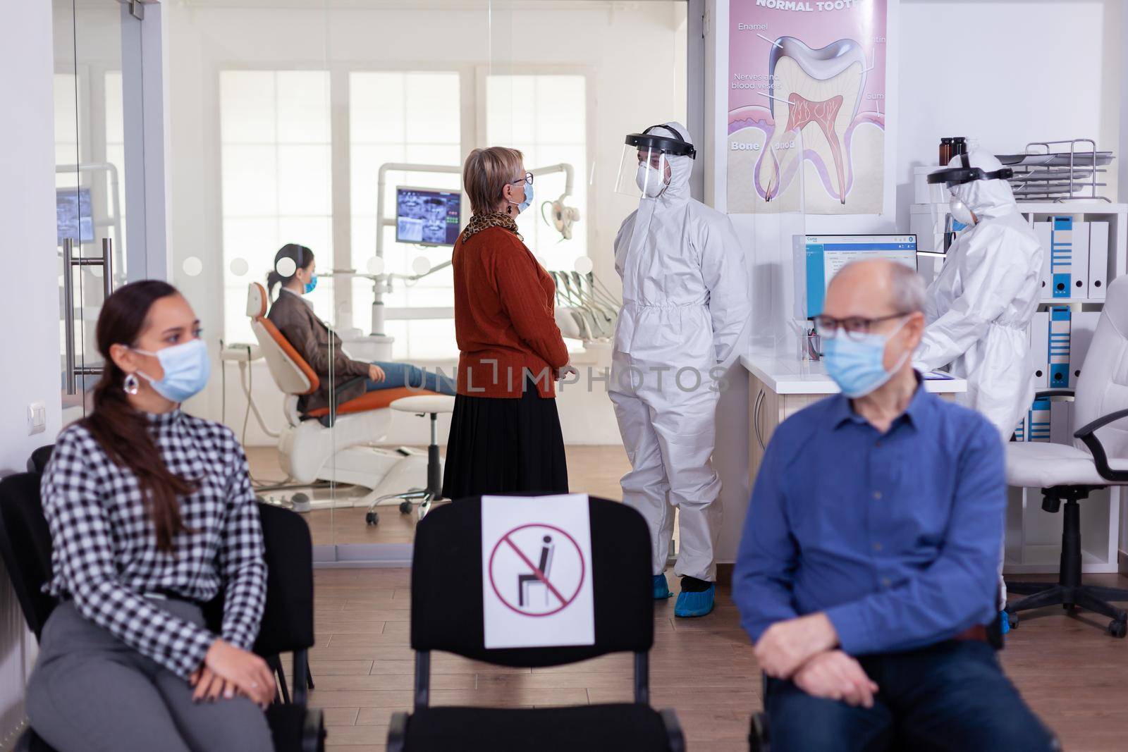 Senior woman with face mask discussing with dentist dressed with ppe suit in stomatology clinic, as safety precaution during global pandemic with coronavirus.