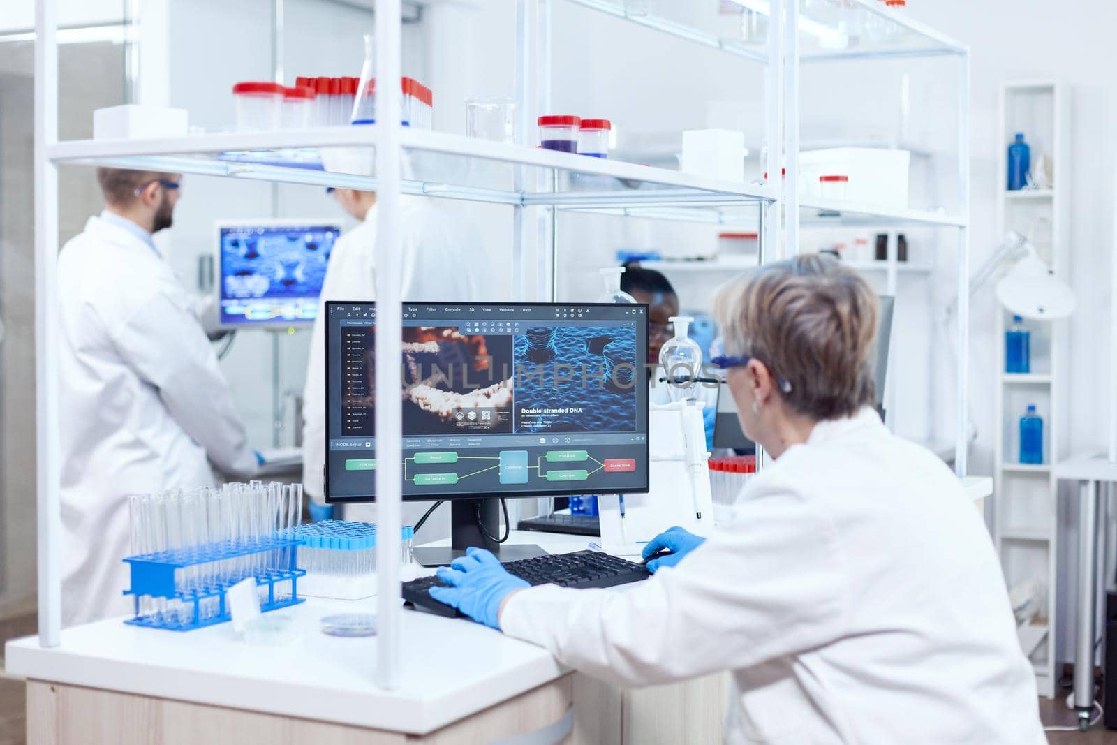 Team of multy ethnical chemist reseachers working together with protective glasses. Senior scientist in pharmaceuticals laboratory doing genetic research wearing lab coat with team in the background.