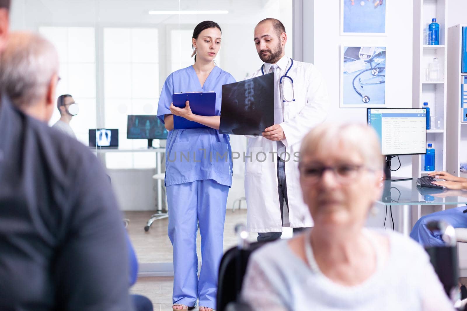 Nurse looking at radiography while talking with doctor about patient diagnosis in hospital waiting area. Invalid old woman in wheelchair. Senior man waiting for medical examination.