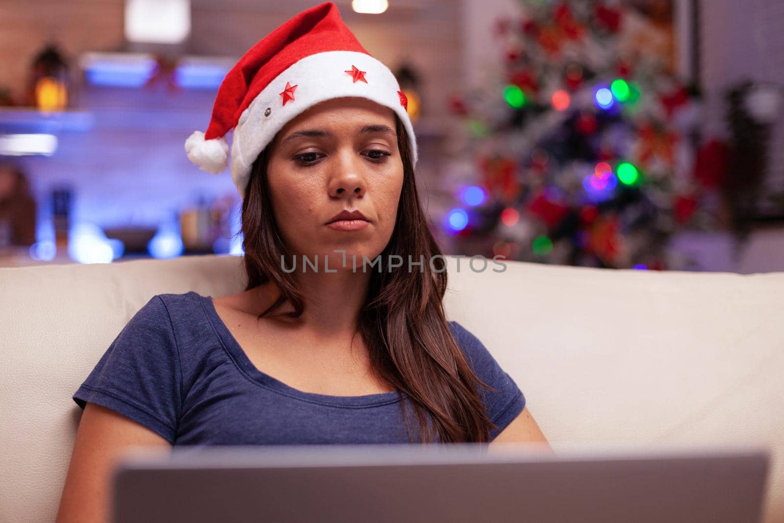 Female adult reading business email on laptop computer working during christmastime sitting comfortable on sofa in xmas decorated kitchen. Woman enjoying winter holiday celebrating christmas season