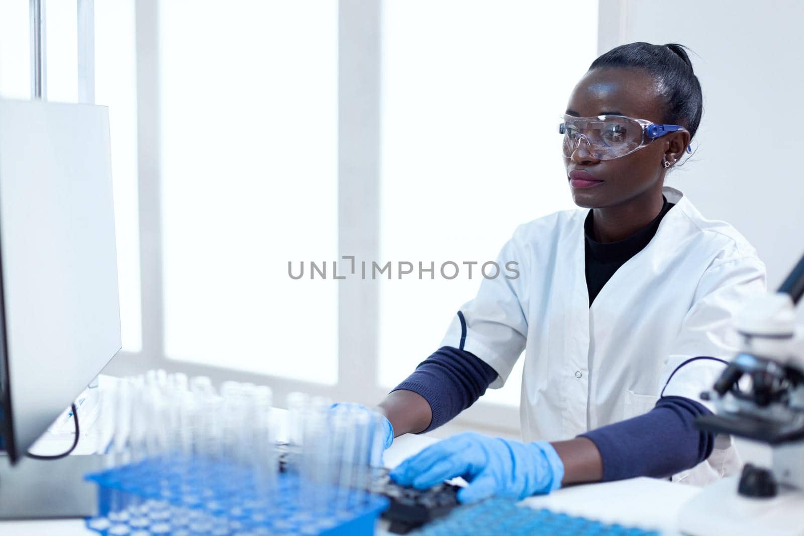 African chemist typing rapport on computer about virus study on computer sitting at her workplace. Black healthcare scientist in biochemistry laboratory wearing sterile equipment.