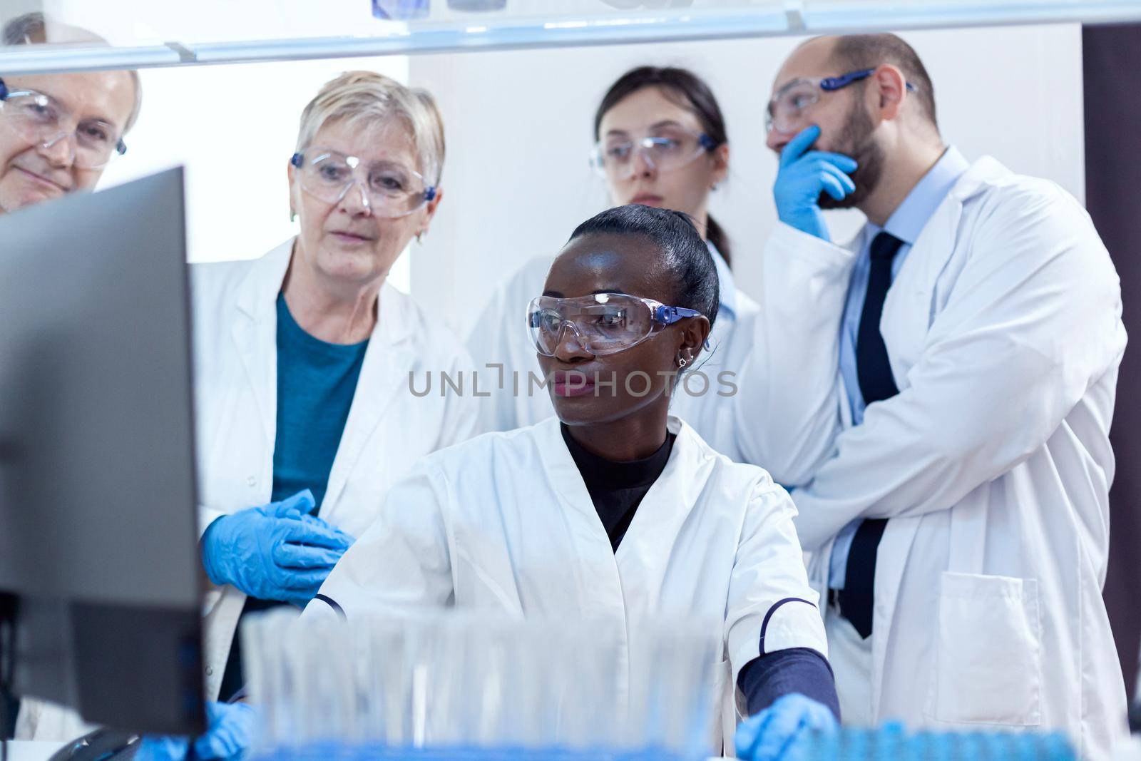 Group of scientists in medicine lab looking at computer display analysing virus. Black healthcare researcher in biochemistry laboratory wearing sterile equipment.