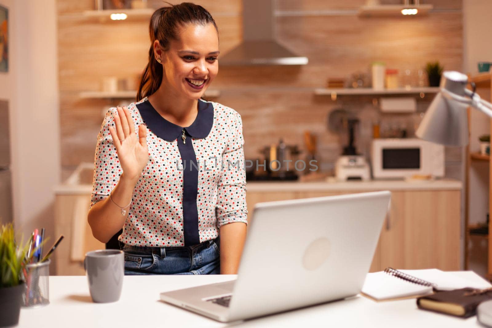 Cheerful woman waving during a video call while working late at night from home kitchen. Employee using modern technology at midnight doing overtime for job, business, career, network, lifestyle.