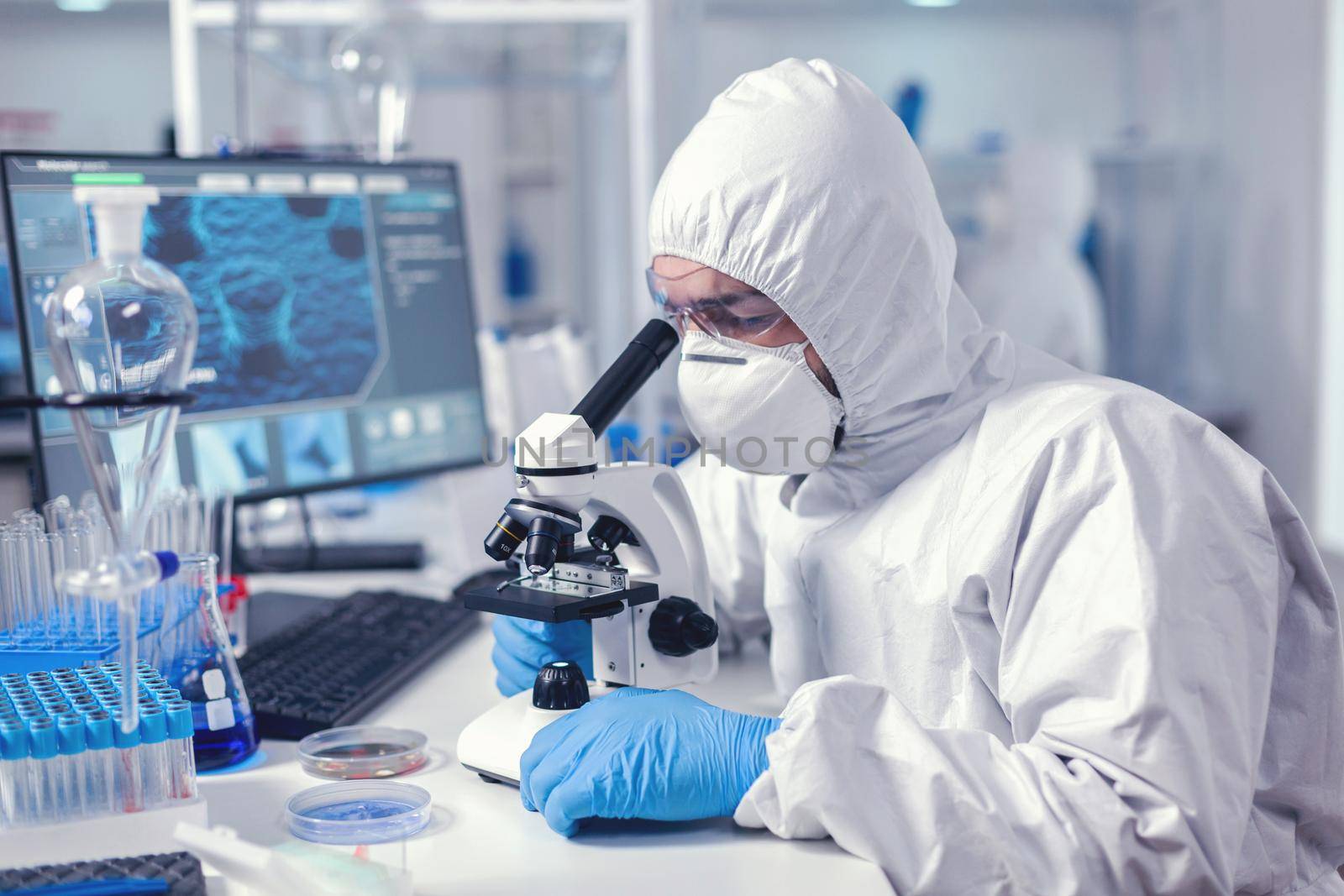 Medic working with microscope analyzing sample using microscope in time of pandemic. Virolog in coverall during coronavirus outbreak conducting healthcare scientific analysis.