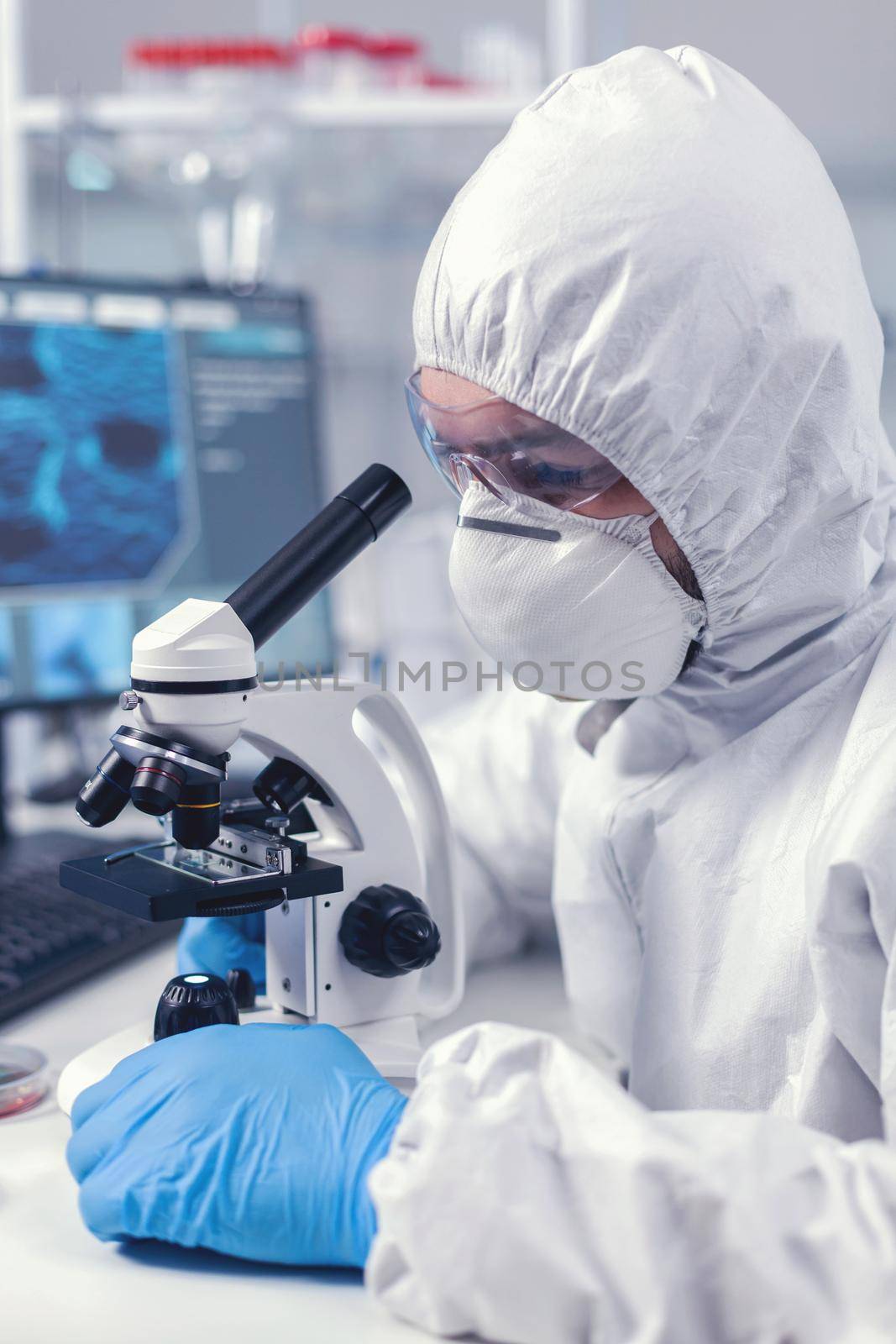 Young scientist look through microscope while doing research in scientific laboratory dressed in ppe suit. Virolog in coverall during coronavirus outbreak conducting healthcare scientific analysis.