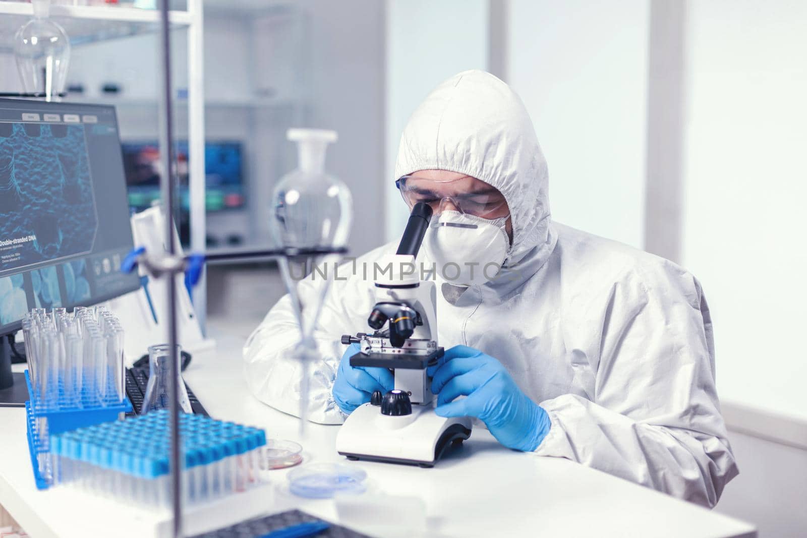 Analyzing virus in microbiology lab using microscope wearing ppe suit and glasses. Virolog in coverall during coronavirus outbreak conducting healthcare scientific analysis.