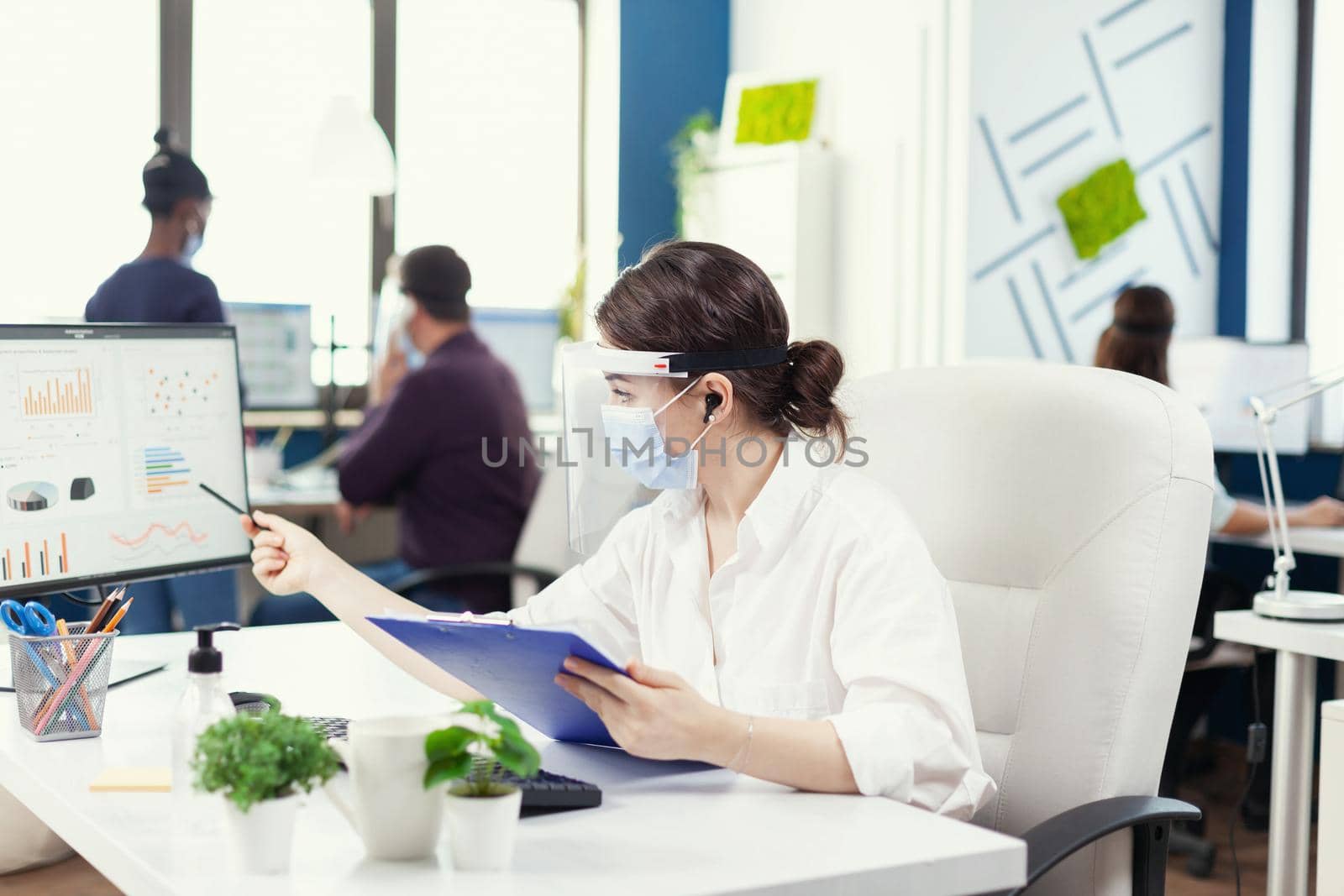 Focused businesswoman on a remote call using wireless headphones wearing face mask against covid19. Employees with visors working in corporate company workspace respecting social distance analysing data and graphs.