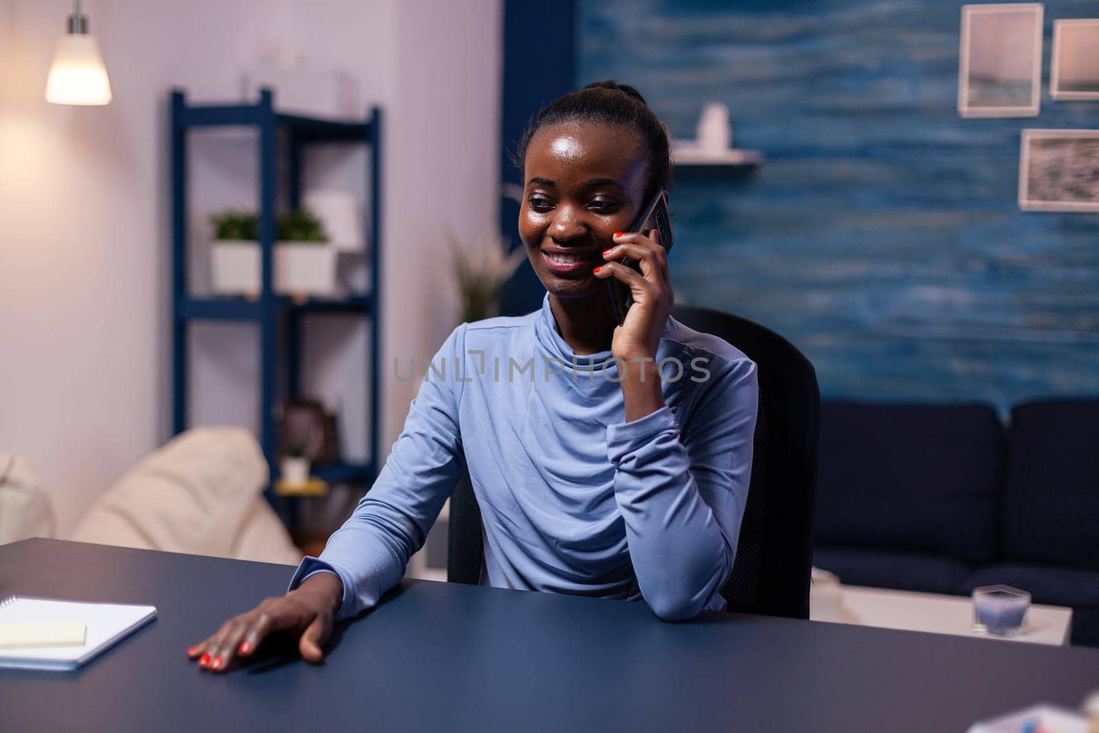 African business woman speaking on smartphone working late at night from home office. Busy focused freelancer using modern technology network wireless doing overtime.