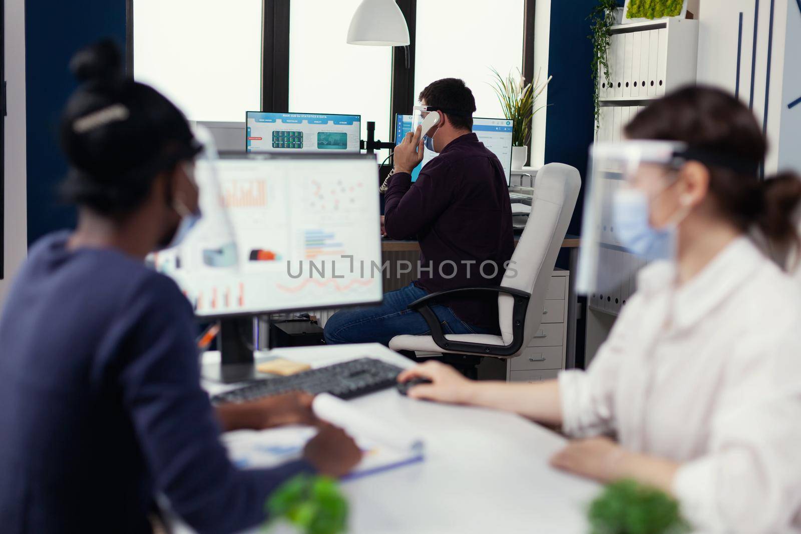 Employee having a phone conversatiion with client sitting at desk wearing face mask for covid19. Multiethnic team working in company with new normal respecting social distance during global pandemic with coronavirus.