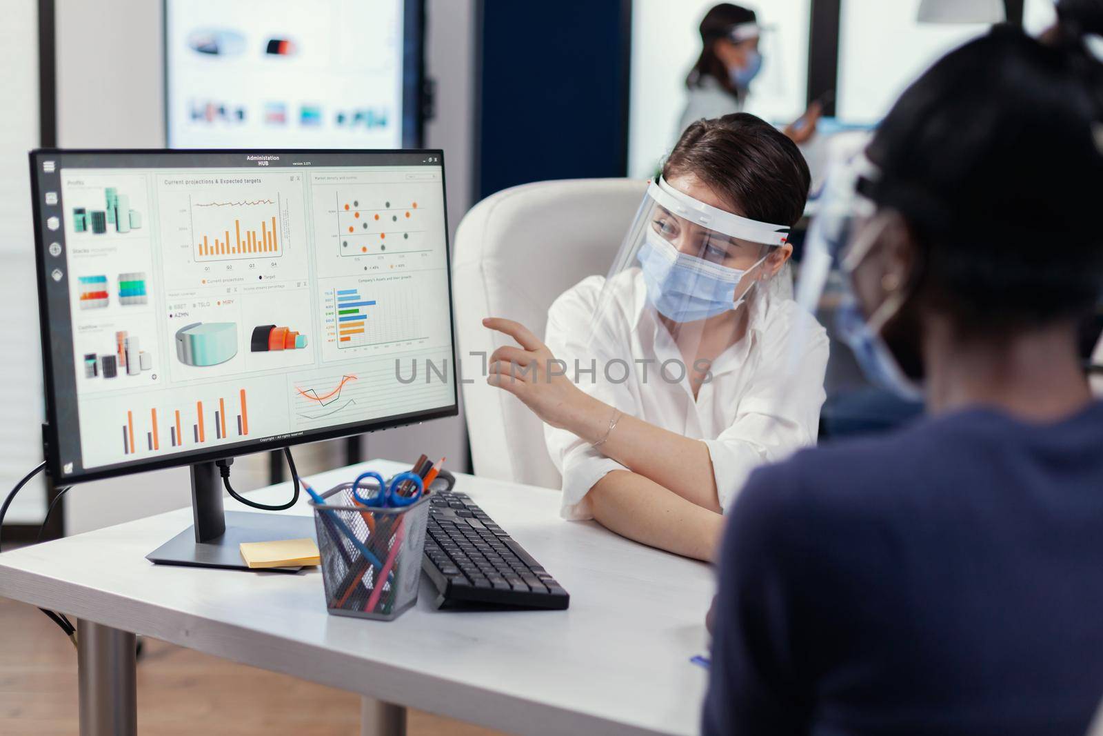 Manager sitting at desk pointing at financial graph during discussion with african emply wearing face mask. Multiethnic team working in company with new normal respecting social distance.