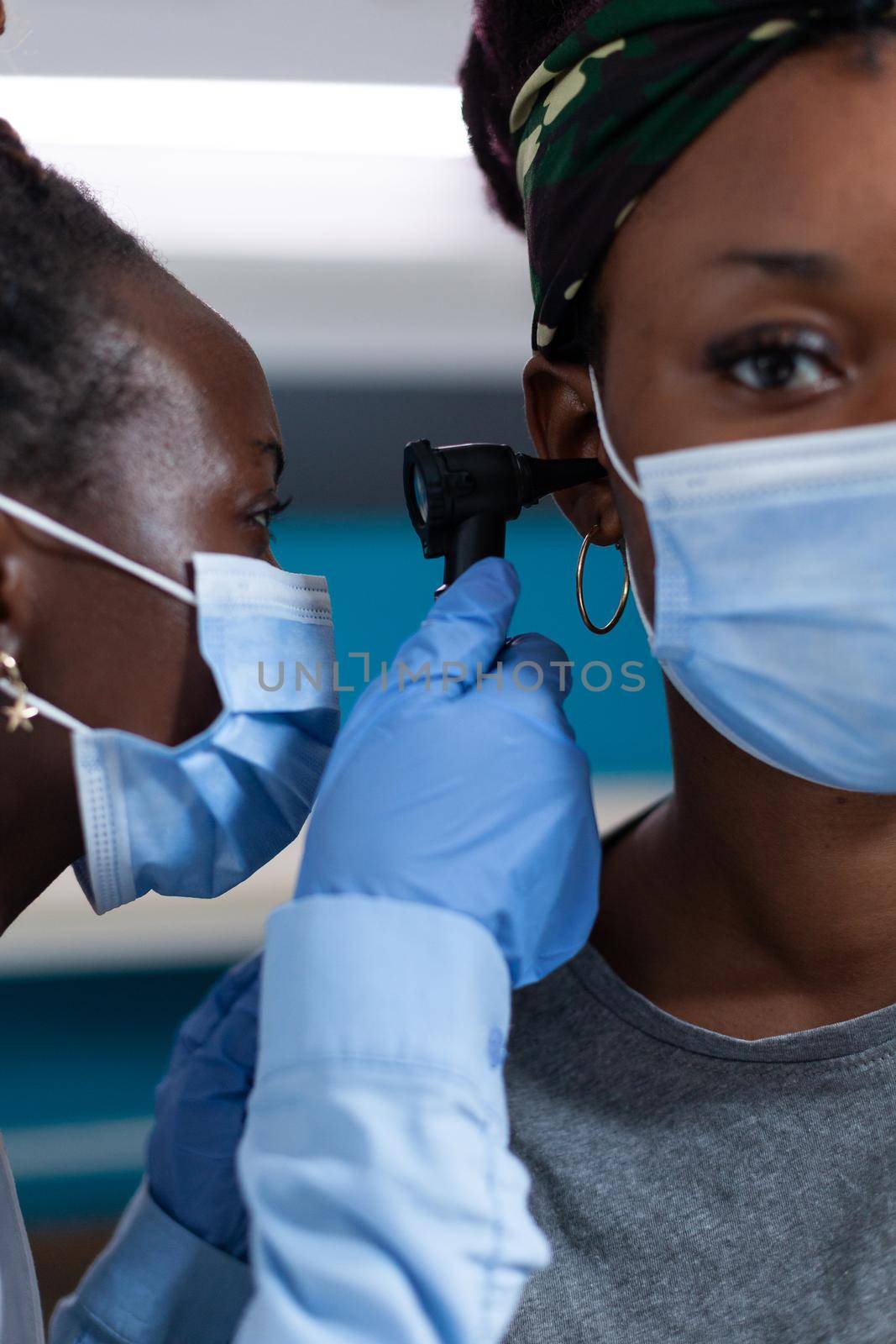 Closeup of african american otologist doctor with protection face mask against covid19 analyzing patient ear using medical otoscope during clinical appointment in hospital office. Otoscopy tool