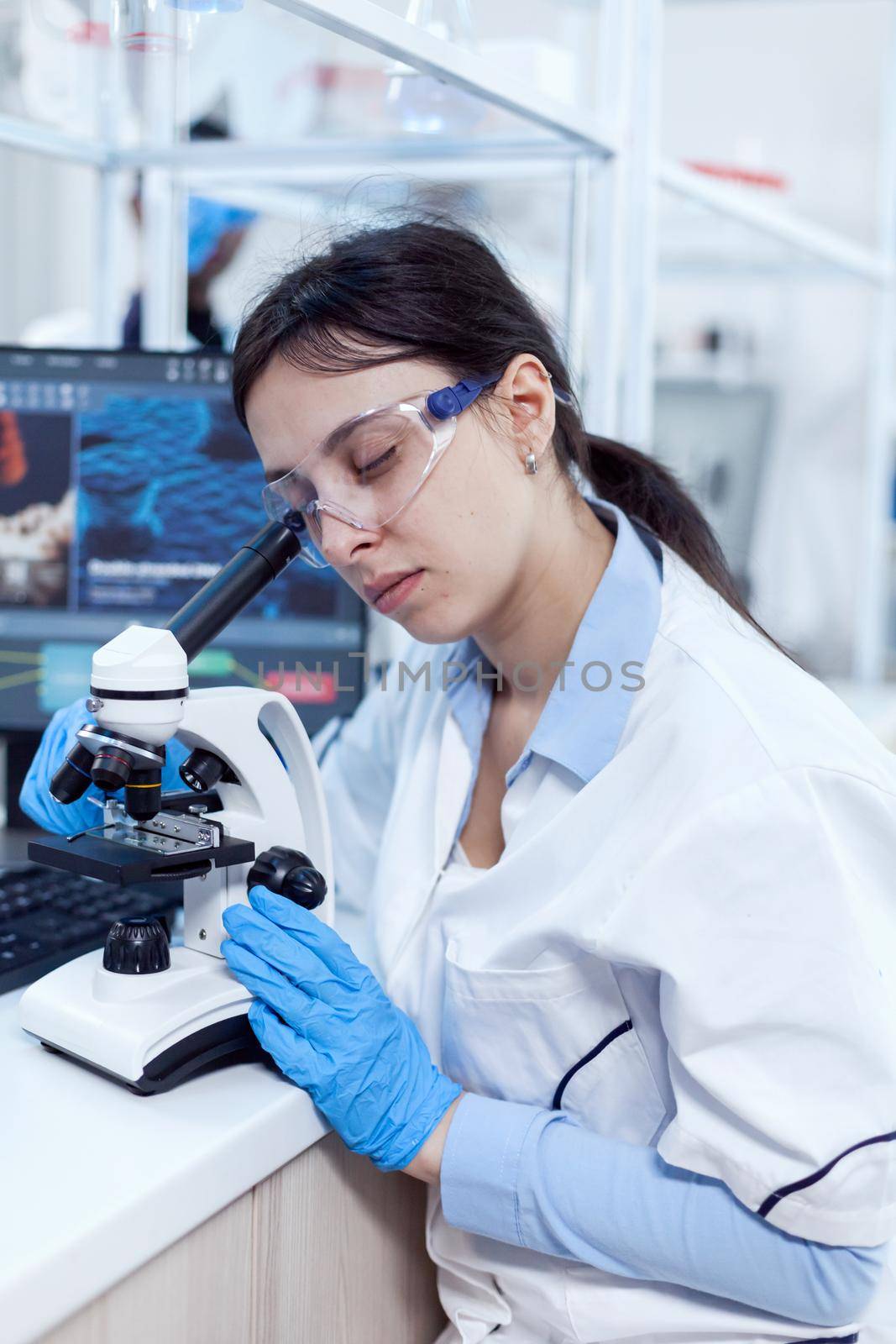 Scientist in protective eyewear doing virus experiment using microscope. Medical technician wearing white coat in sterile laboratory doing solution analysis.
