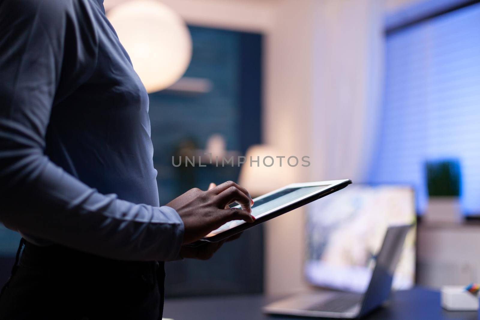 Overworked african woman holding tablet pc from home office in the evening. Busy focused employee using modern technology network wireless doing overtime writing.