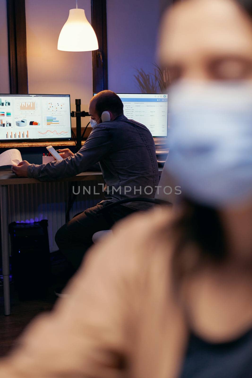 Businessman with face mask working on online business discussing with clients on the phone. Woman following social distancing rules due to coroanvirus pandemic while working late hours at the office.