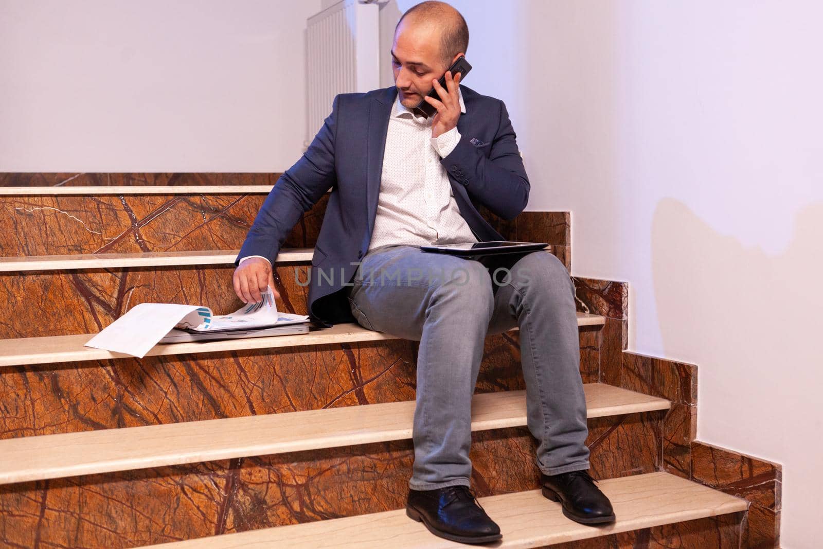 Exhausted overworked businessman talking on phone with team about project deadline.Entrepreneur working late in evening corporate sitting on stairs in building office.
