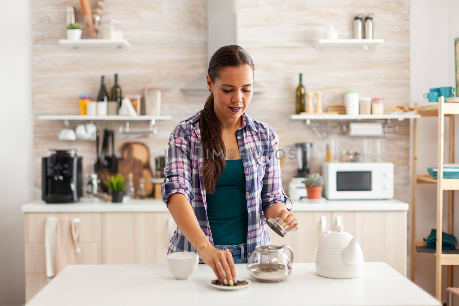 Woman preparing green tea for breakfast in kitchen. Preparing tea in the morning, in a modern home sitting near the table. Putting with hands, healthy herbal in pot.