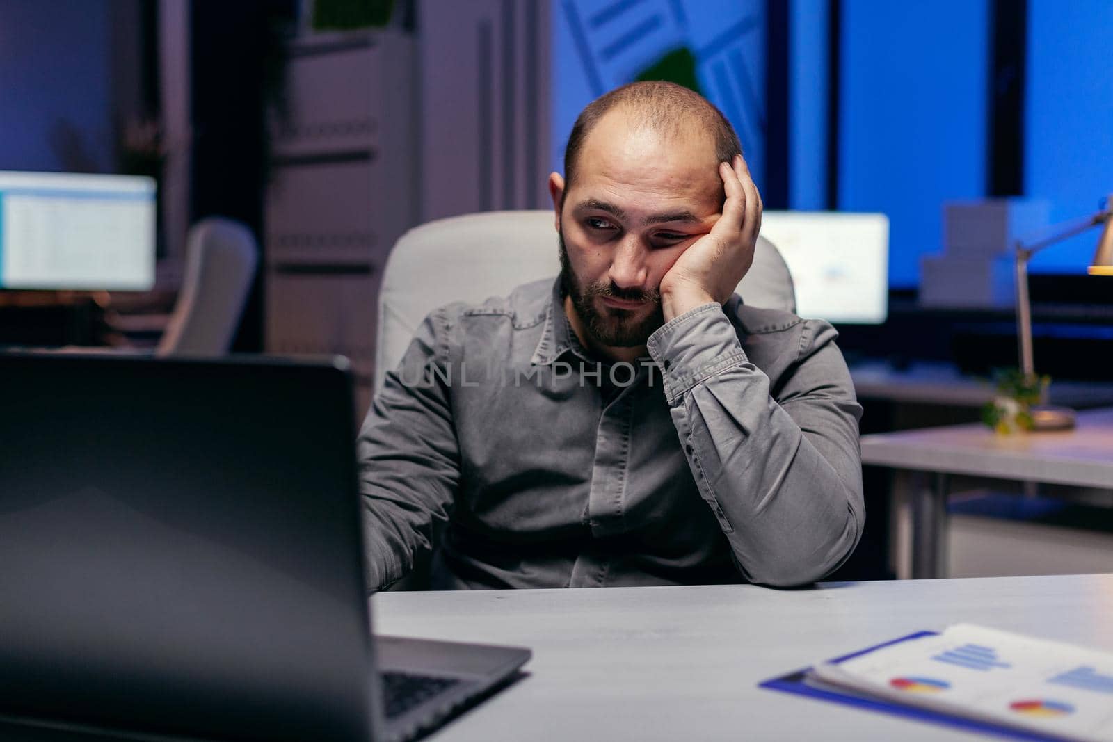 Tired businessman sit at the computerin the evening works on deadline. Workaholic employee falling asleep because of working late at night alone in the office for important company project.