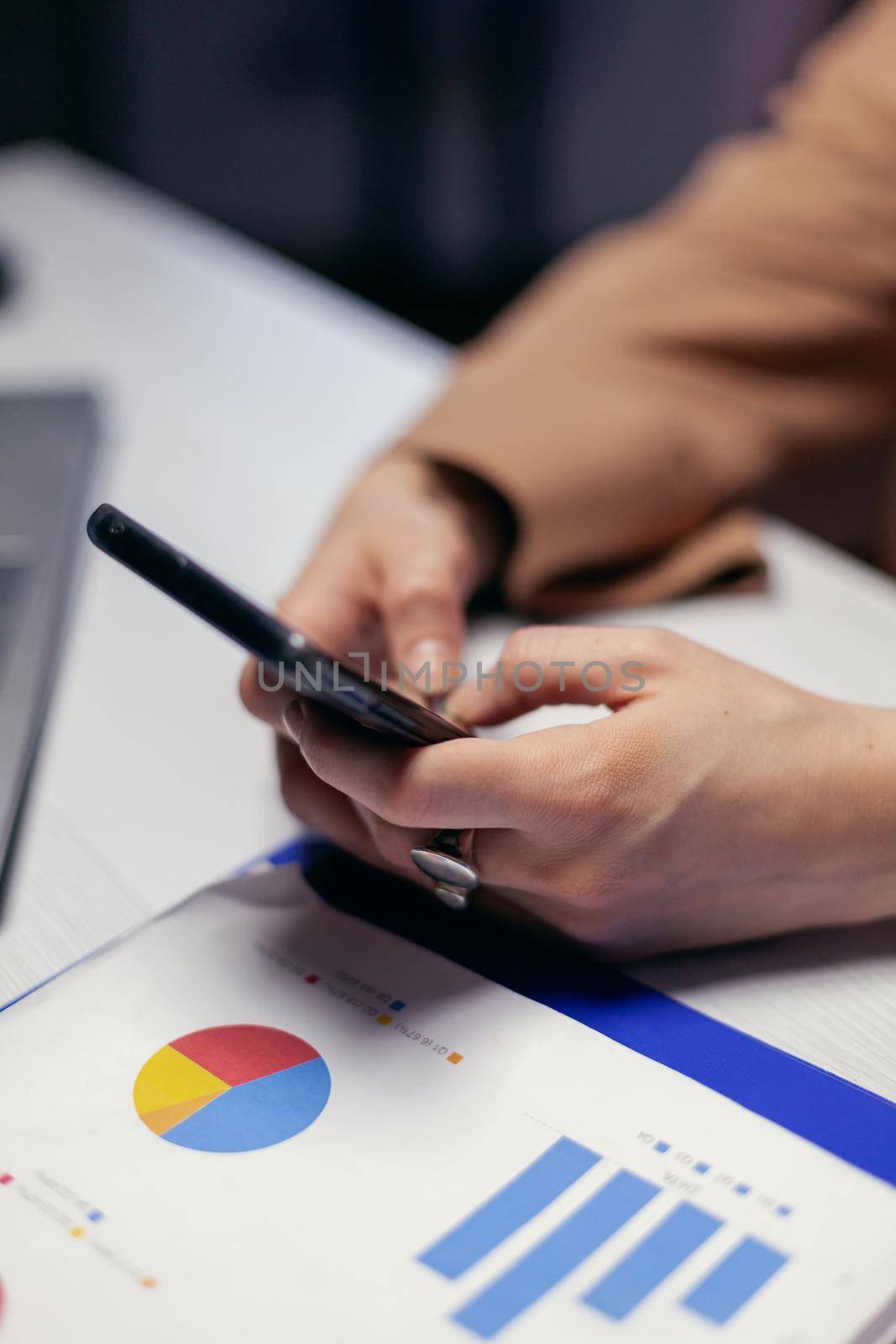 Close up of entrepreneur sending sms holding phone sitting at workplace in the evening. Businesswoman texting late at night while working on important project using smartphone .