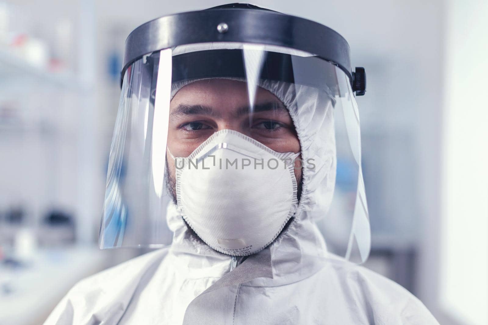 Tired healthcare scientist loooking at camera wearing ppe suit with face shieldin lab. Overworked researcher dressed in protective suit against invection with coronavirus during global epidemic.