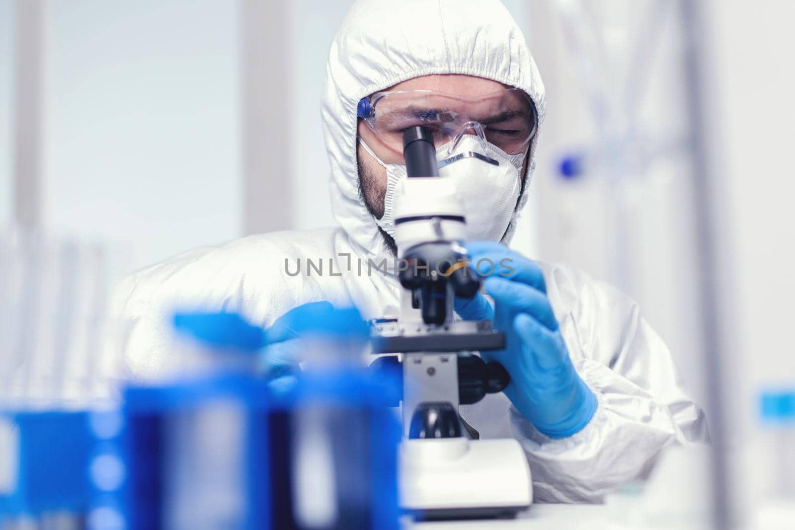 Microbiologist researcher wearing ppe and protection glasses in medicine lab looking through microscope. Scientist in protective suit sitting at workplace using modern medical technology during global epidemic.