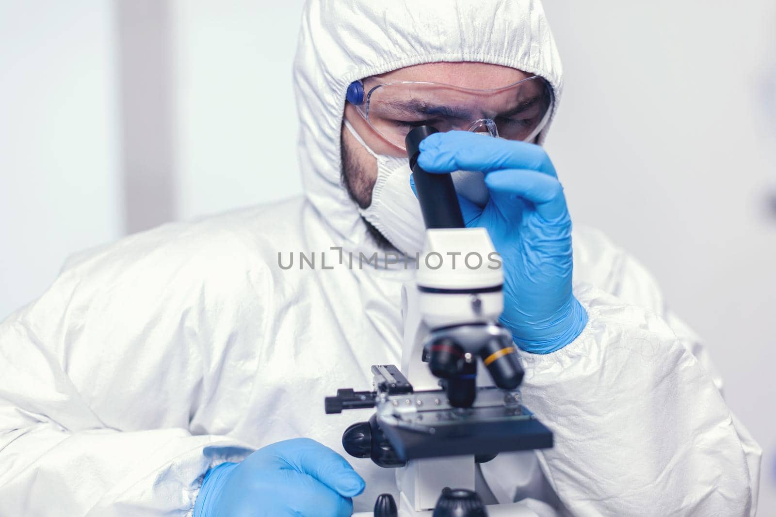 Medical researcher looking throug microscope at virus sample dressed in ppe suit. Scientist in protective suit sitting at workplace using modern medical technology during global epidemic.